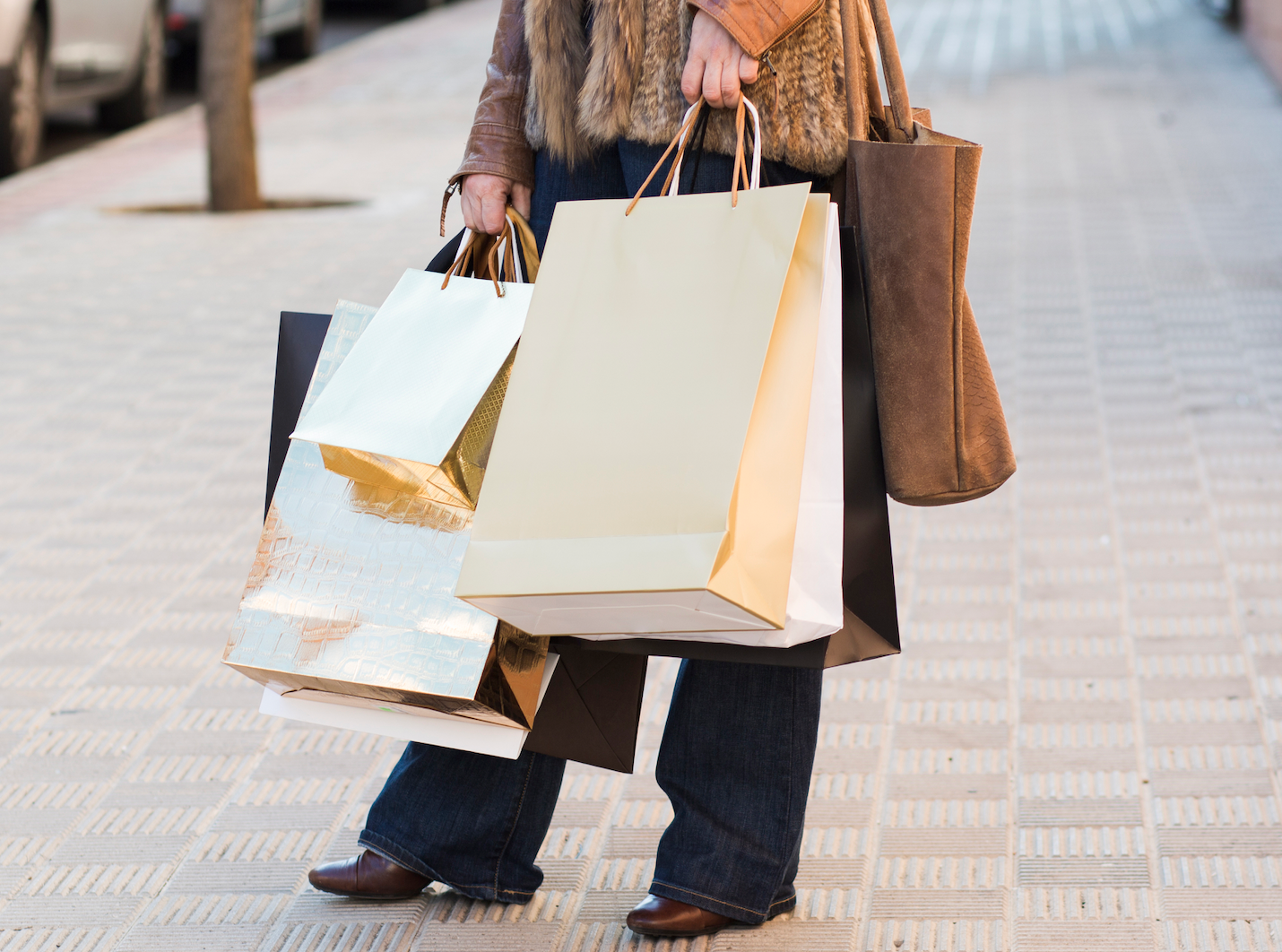A person walking on a paved sidewalk carrying multiple paper shopping bags and a large brown tote bag.