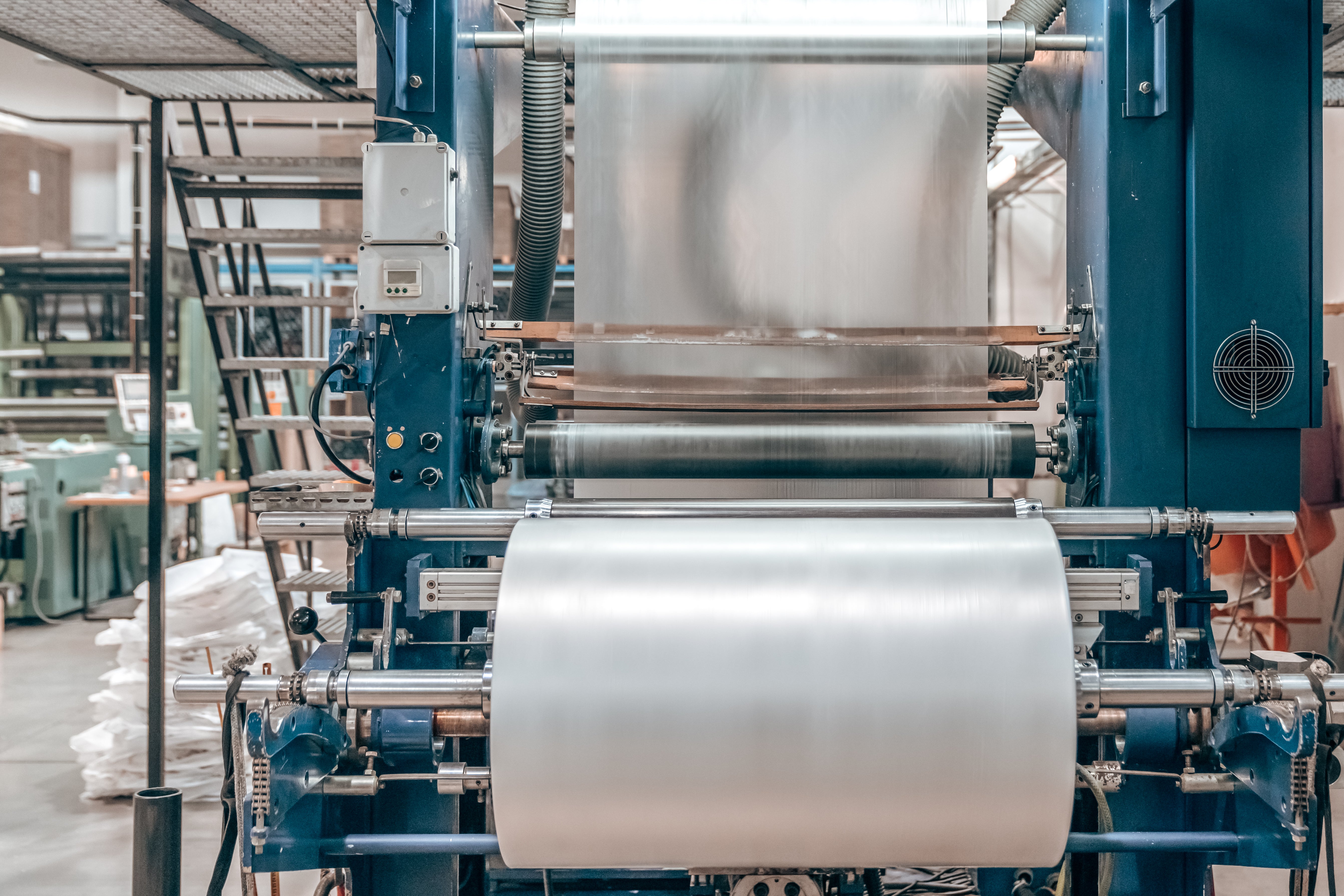 A large industrial roll of translucent white plastic film mounted on a blue metal processing machine inside a factory setting